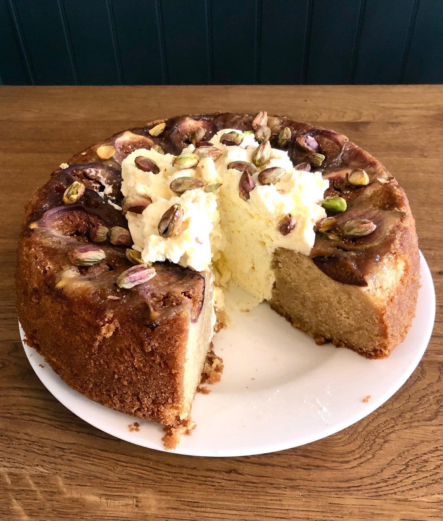 A whole cake photographed by Petit Fi, with a slice removed, cream and a scattered topping on its plate
